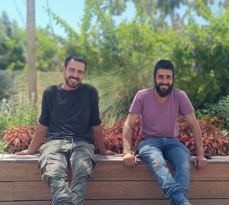 שני גברים מחייכים יושבים על ספסל עץ כשברקע צמחיה שופעת, המבטים שלהם פונים למצלמה